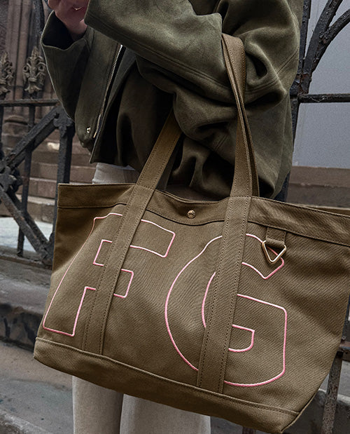 A person wearing an olive green jacket carries a large olive green canvas tote bag with pink outlined lettering on a city street.