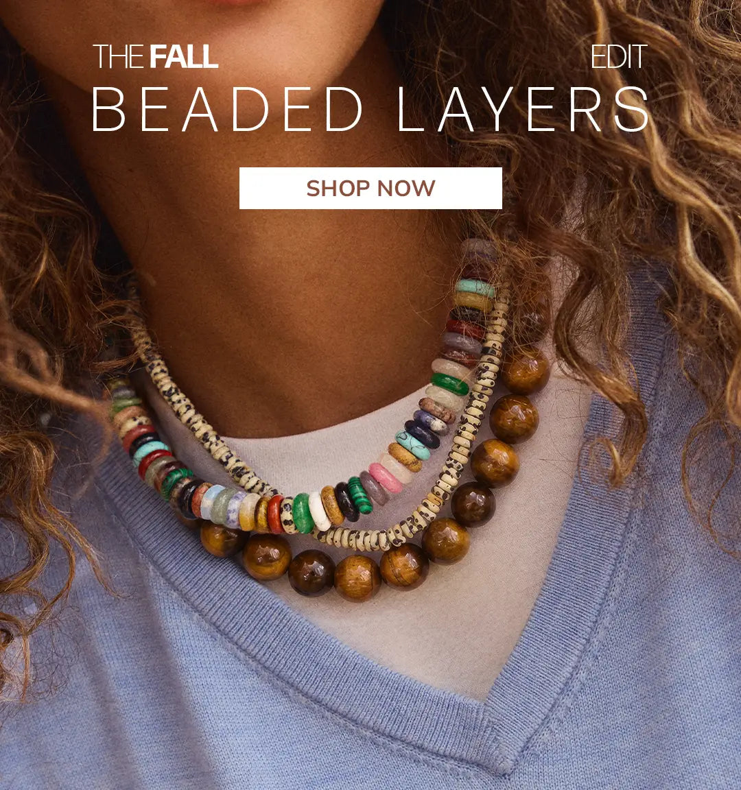 An editorial shot of a model wearing three beaded necklaces with a light blue sweater layered on top of a white tshirt.