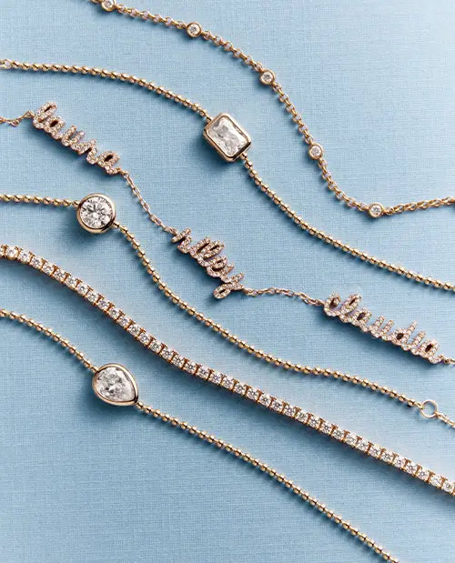 Six personalized gold name bracelets with clear gemstones are arranged diagonally on a textured light blue background. The nameplates include 'laura', 'riley', 'chloe', and three stone-set names.