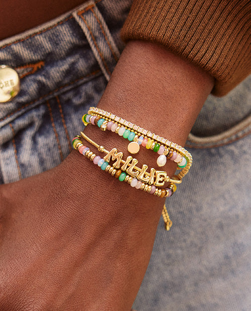 Close-up of layered gold bracelets featuring a personalized name bracelet, colorful pastel beads, gold charms, and a sparkling crystal tennis bracelet.