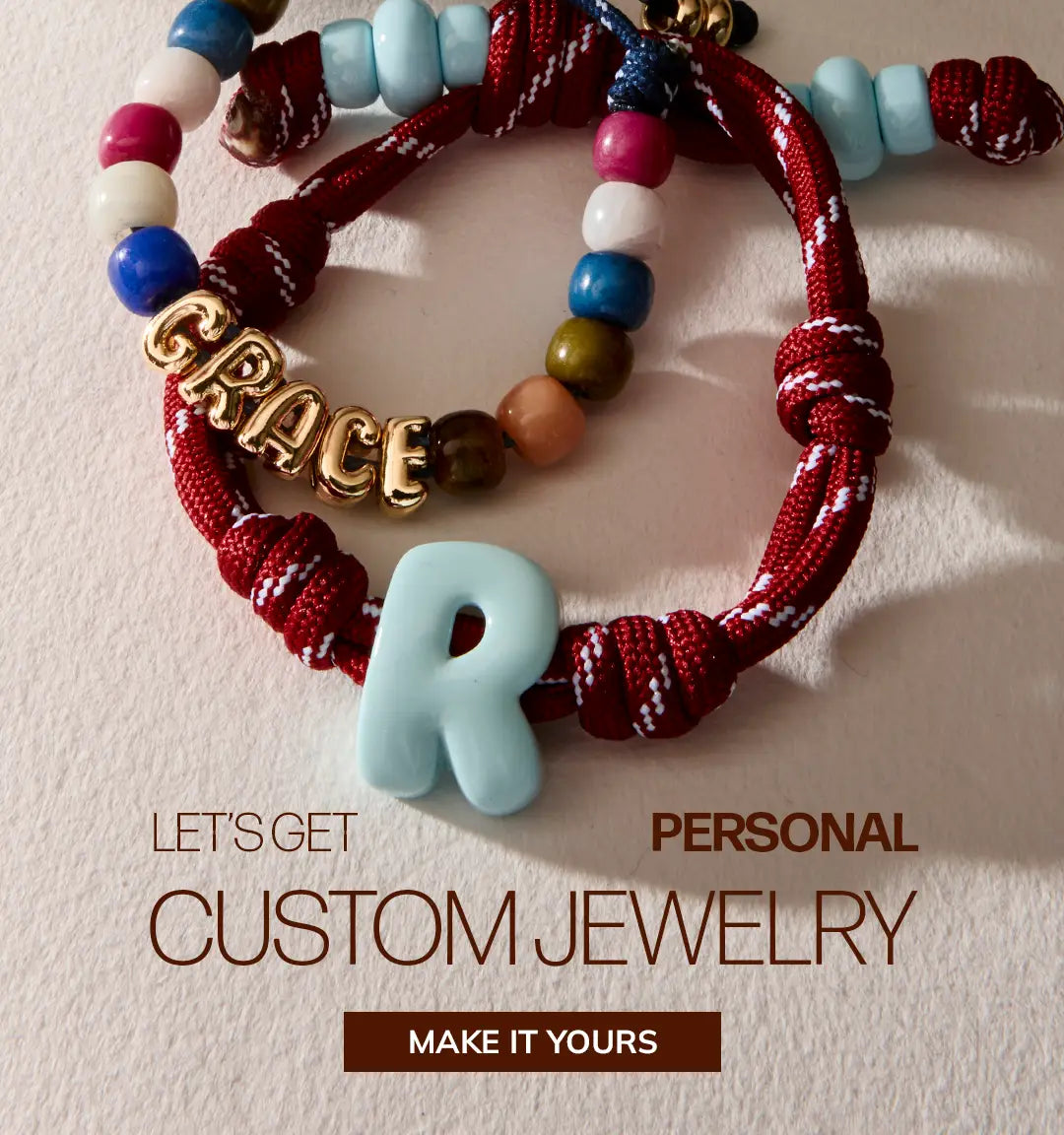 Red rope bracelet with a light blue “R” and a beaded “GRACE” bracelet. Text reads “Let’s get personal: Custom Jewelry.” Button: “Make It Yours.”