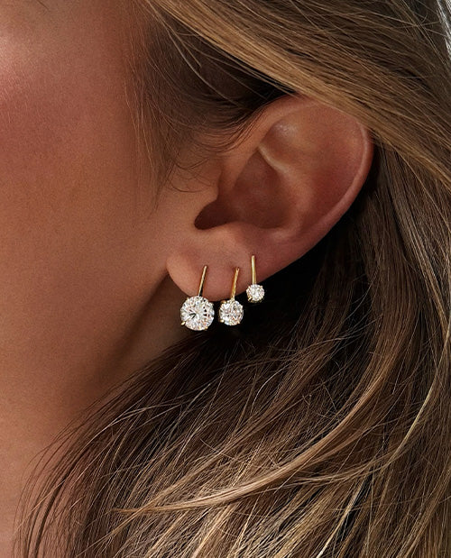Close-up of three sparkling clear crystal drop earrings in graduated sizes, featuring gold-tone settings styled in multiple ear piercings.