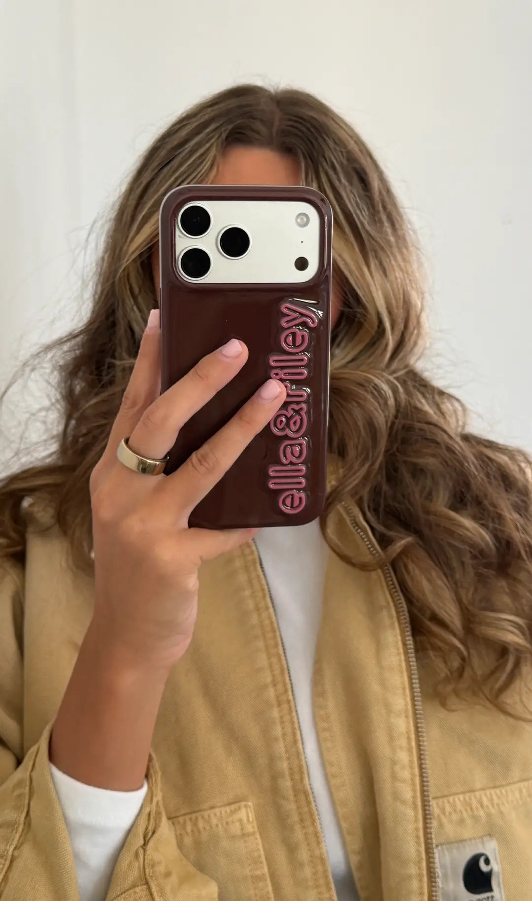 Woman holds a phone in a burgundy case with raised pink lettering reading “ella&riley” in front of her face.