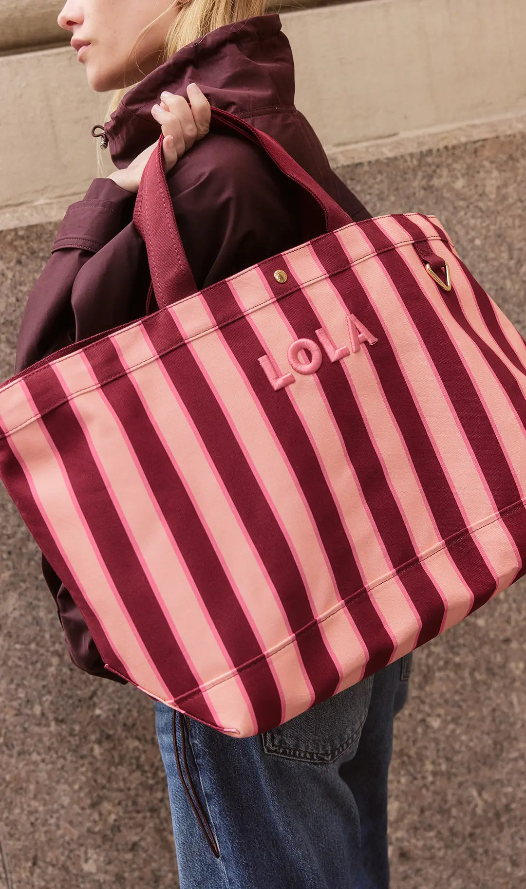 Woman carries a burgundy-and-pink striped canvas tote embroidered “LOLA” over her shoulder.