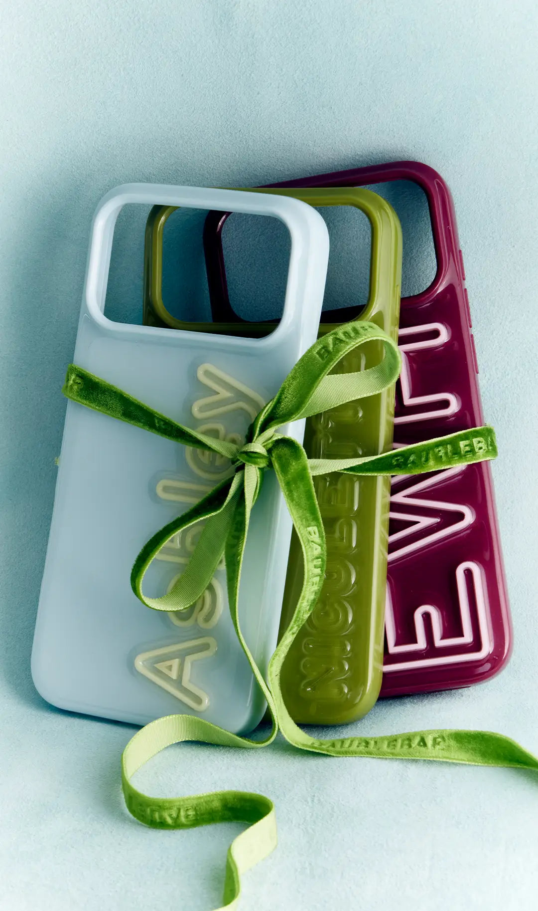 Three personalized phone cases in light blue, olive and burgundy with raised name lettering, tied with a green velvet bow.