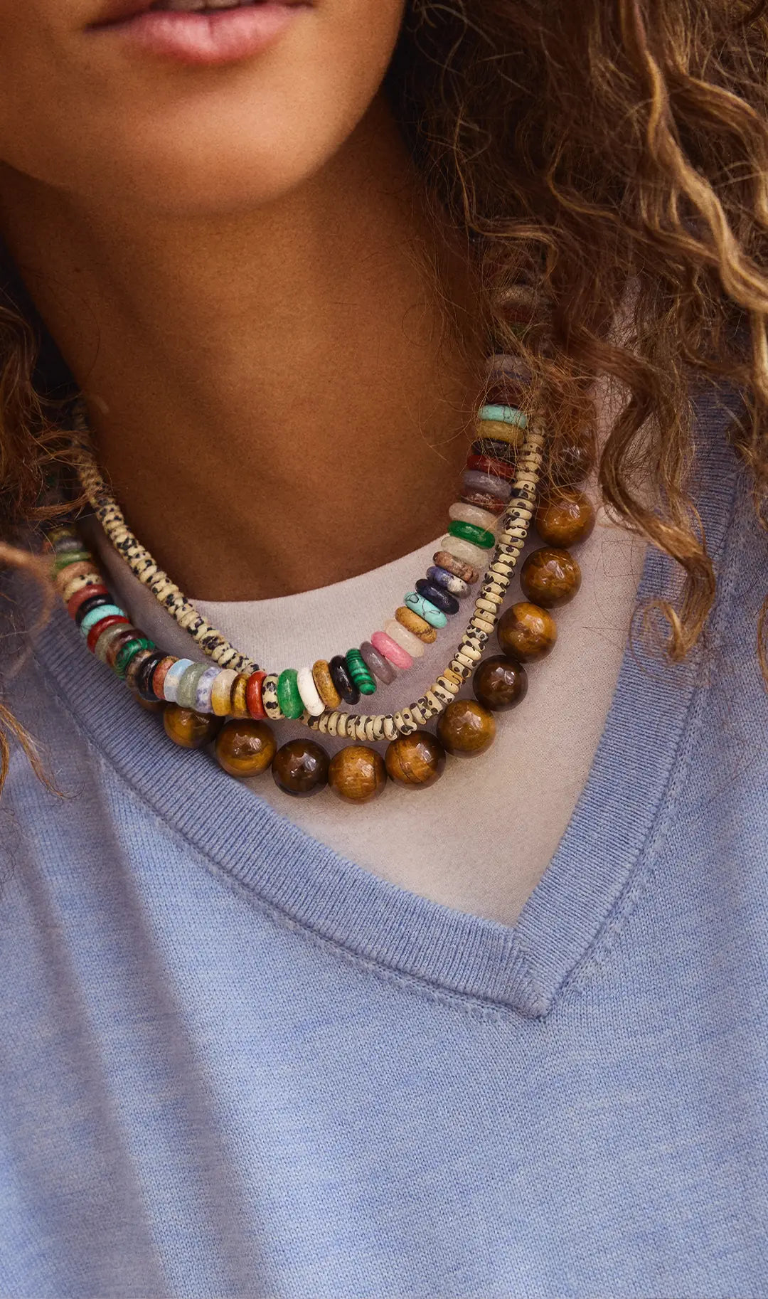 Woman in a blue V-neck sweater wears layered necklaces of multicolor stone discs, speckled beads and large tiger’s-eye beads.
