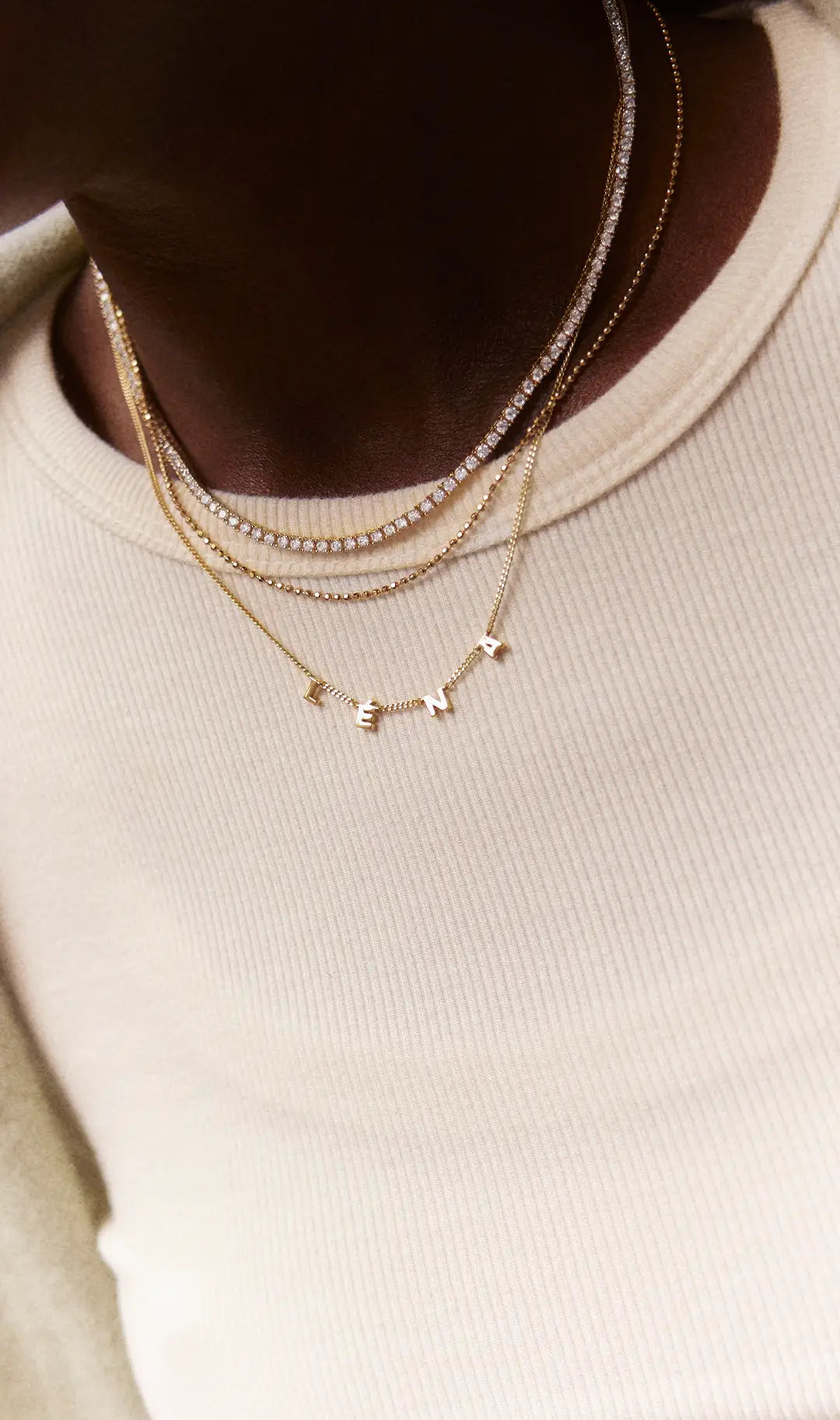 Woman in a cream ribbed top wears layered gold chains, a crystal tennis necklace and a letter necklace spelling “LENA.”