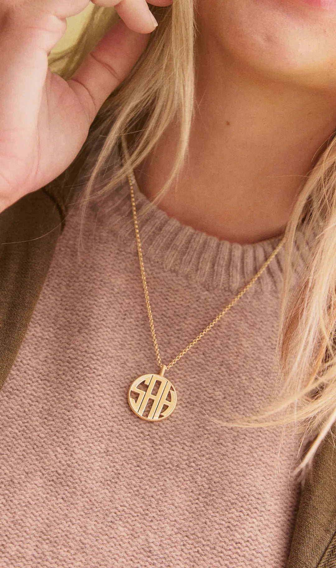 Woman in a pink knit sweater wears a round gold pendant with three block-letter initials "SAH" on a thin chain.