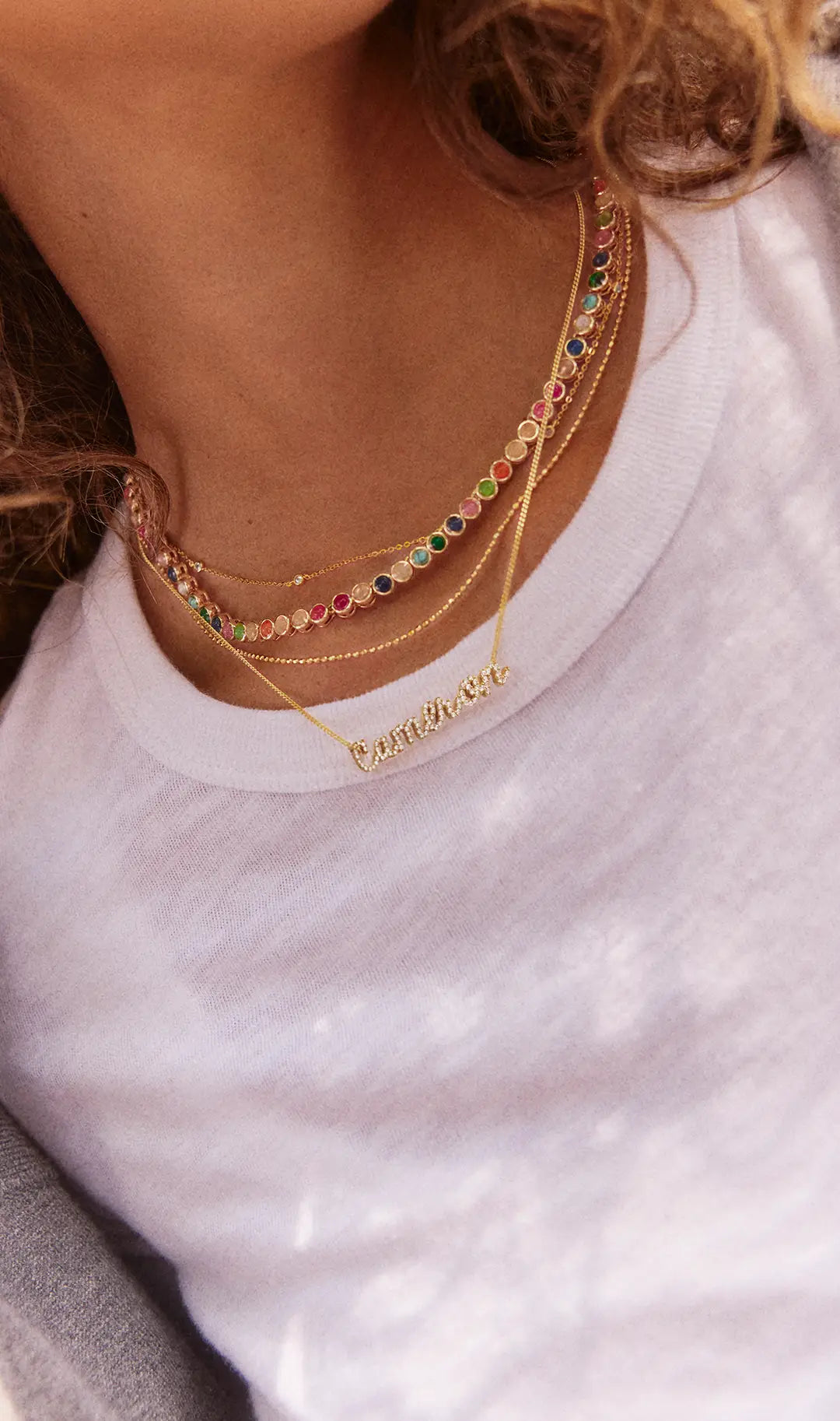 Woman in a white tee wears layered necklaces: a rainbow gemstone tennis necklace, fine gold chains and a crystal script name pendant "Cameron."