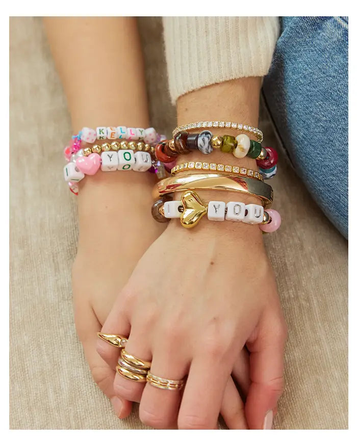 Clasped hands wearing stacked beaded letter bracelets reading “KELLY” and “I ♥ YOU” with gold bangles and rings.