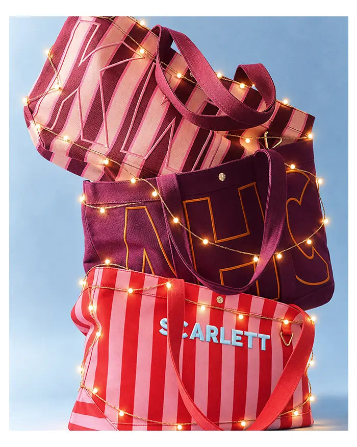 Three stacked monogrammed canvas totes wrapped in twinkle lights, the red-and-pink striped one reading “SCARLETT.”
