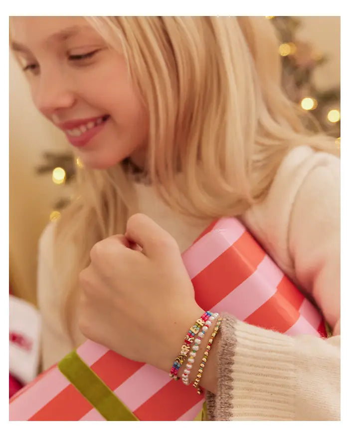 Smiling girl holding a striped gift wears three stacked bracelets: beaded letters, pearls and gold beads.