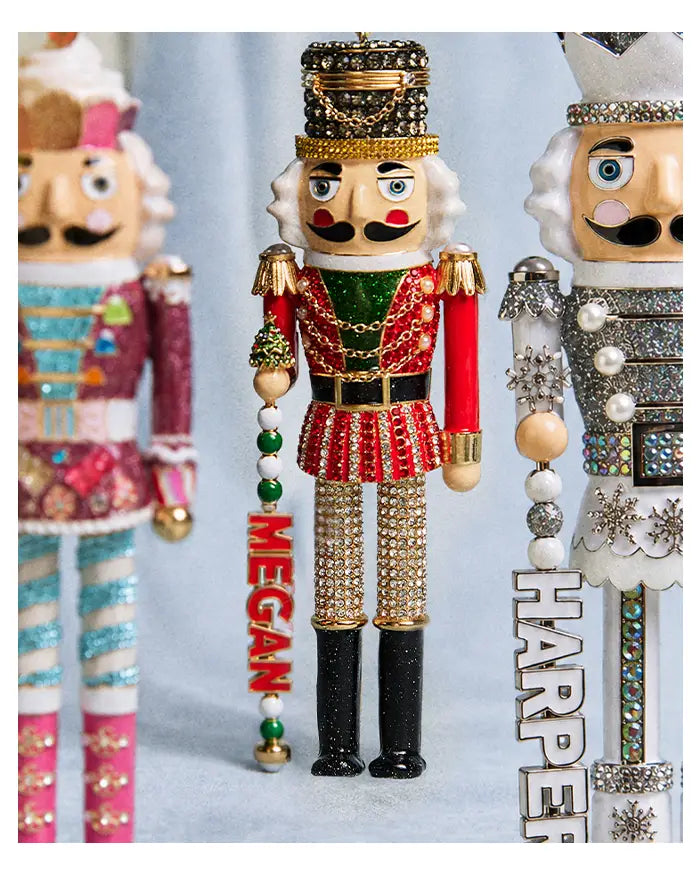 Jeweled red-and-green nutcracker ornament with a “MEGAN” name charm, beside other nutcrackers including one with “HARPER.”