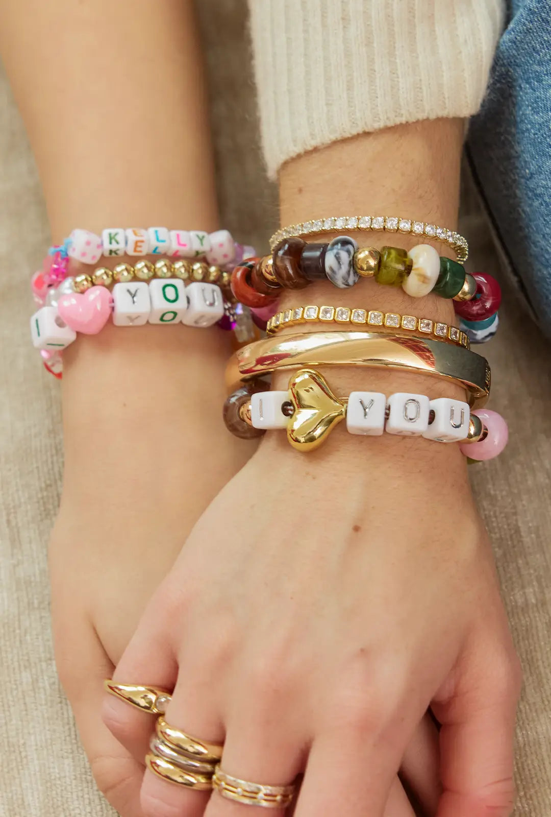 Clasped hands wearing stacked beaded letter bracelets reading “KELLY” and “I ♥ YOU” with gold bangles and rings.