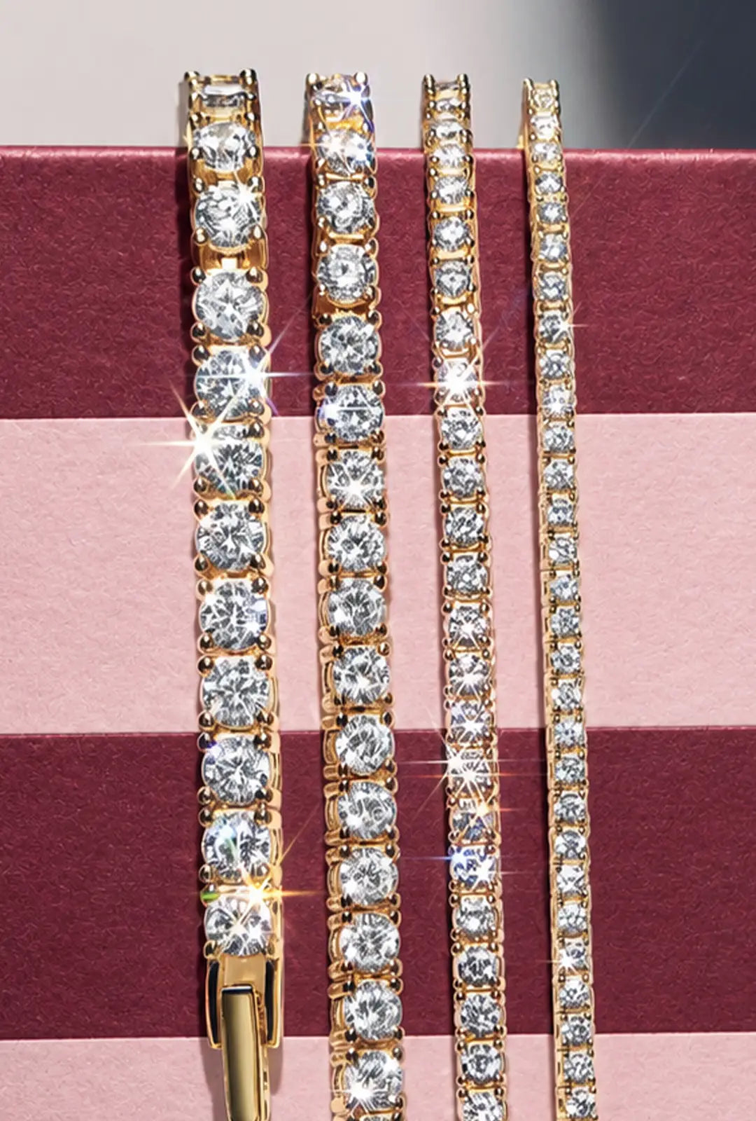 Four gold crystal tennis bracelets in graduated widths hang against a burgundy-and-pink striped box.