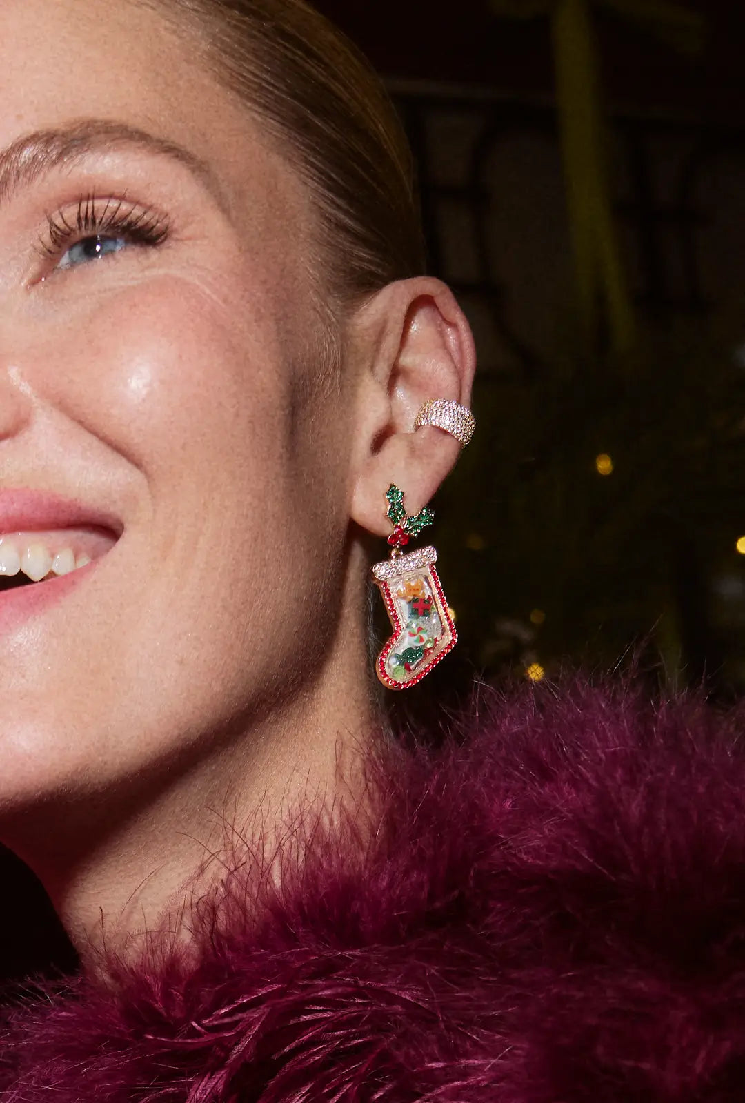 Smiling woman in a plum feather wrap wears a crystal ear cuff and a beaded Christmas stocking drop earring with holly.