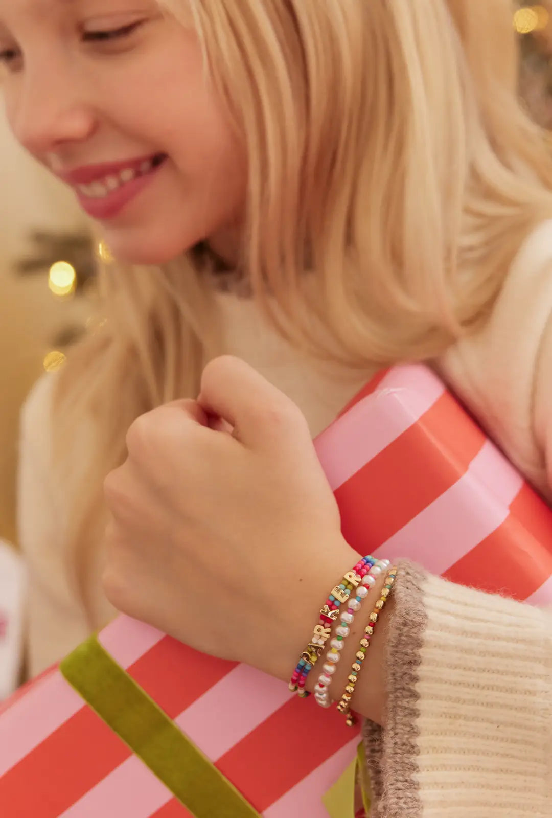 Smiling girl holding a striped gift wears three stacked bracelets: beaded letters, pearls and gold beads.