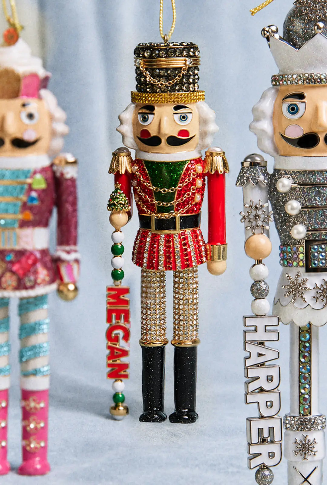 Jeweled red-and-green nutcracker ornament with a “MEGAN” name charm, beside other nutcrackers including one with “HARPER.”