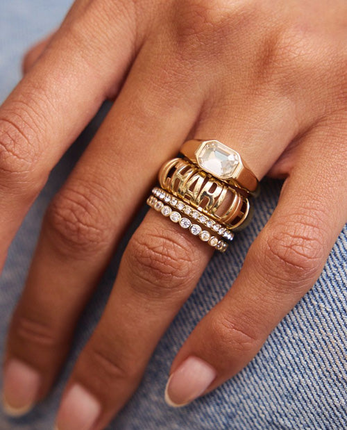 A close-up of a hand wearing a stack of gold rings, including a textured band, a diamond-studded band, and a signet-style ring, resting on blue denim fabric.