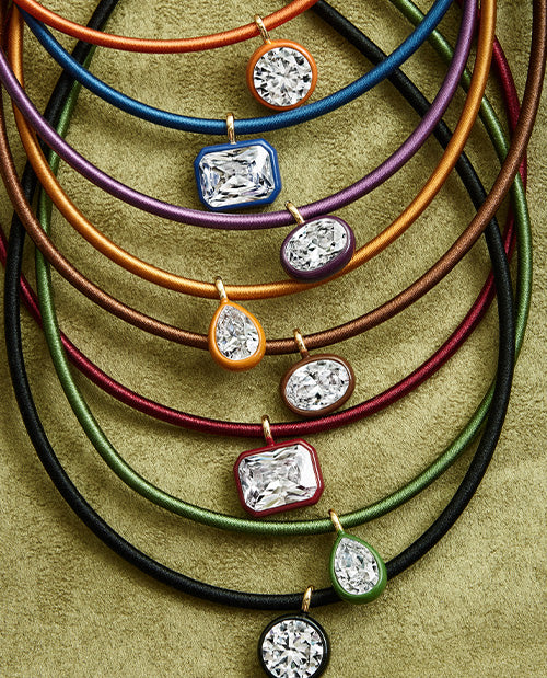 A close-up studio shot of multiple thin cord necklaces arranged in overlapping rows on a textured olive background. Each necklace has a small colored bezel with a clear faceted gemstone pendant, including orange, blue, purple, gold, red, green, and black cords.