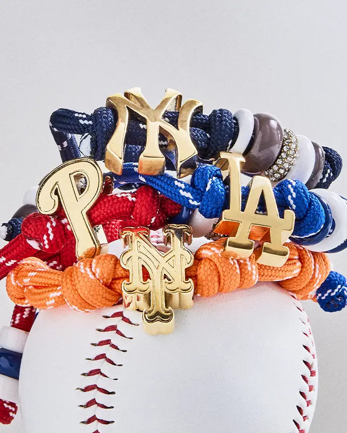 Stacked knotted cord bracelets in navy, blue, red and orange with gold NY, P and LA team logo charms, resting on a baseball.