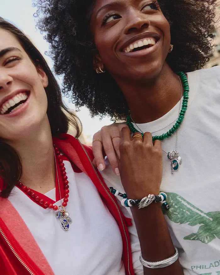 Two laughing friends wear football-themed beaded jewelry: a red bead necklace with charms and a green bead necklace with a wing-charm bracelet.