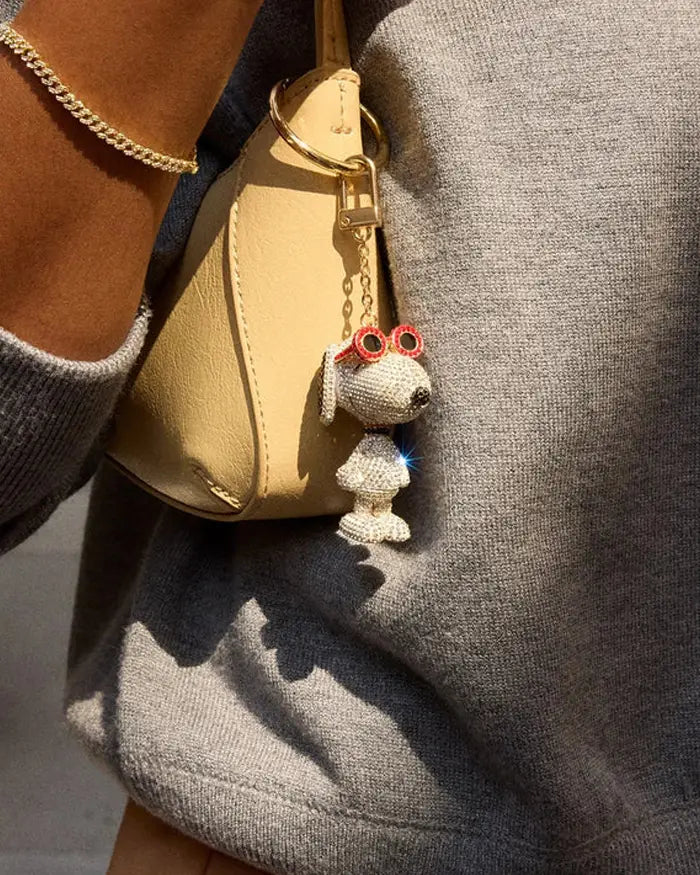 Crystal-covered Snoopy charm in red sunglasses hangs from the strap of a tan shoulder bag.