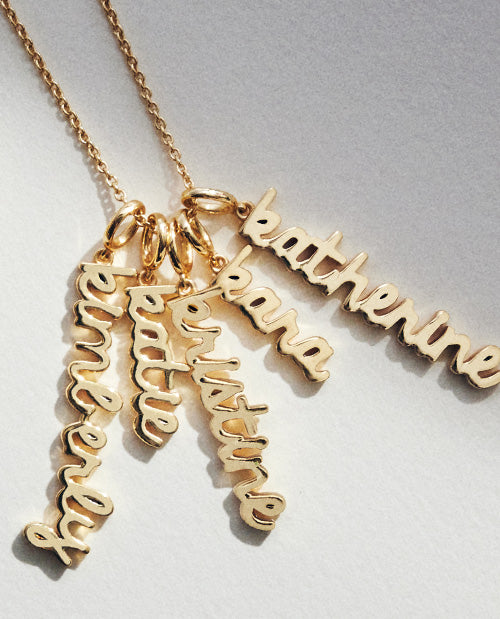 Close-up of gold personalized name necklace charms in a cursive script font, featuring multiple names layered together on a delicate gold chain.